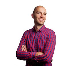 Uśmiechnięty, łysy mężczyzna w czerwono-niebieskiej koszuli w kratę, z założonymi rękami na białym tle. Portret.