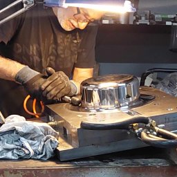 INNOVATICA SPÓŁKA Z OGRANICZONĄ ODPOWIEDZIALNOŚCIĄ - Pracownik w rękawicach poleruje metalowy element na warsztacie. Widoczne narzędzia i lampa oświetlająca. Detale precyzyjnej obróbki metalu w bliskim ujęciu.