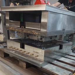 INNOVATICA SPÓŁKA Z OGRANICZONĄ ODPOWIEDZIALNOŚCIĄ - Ciężka, metalowa forma w trakcie montażu z pomocą suwnicy i magnesu, obsługiwana przez mężczyznę w warsztacie. Widoczne elementy hydrauliczne i konstrukcyjne.