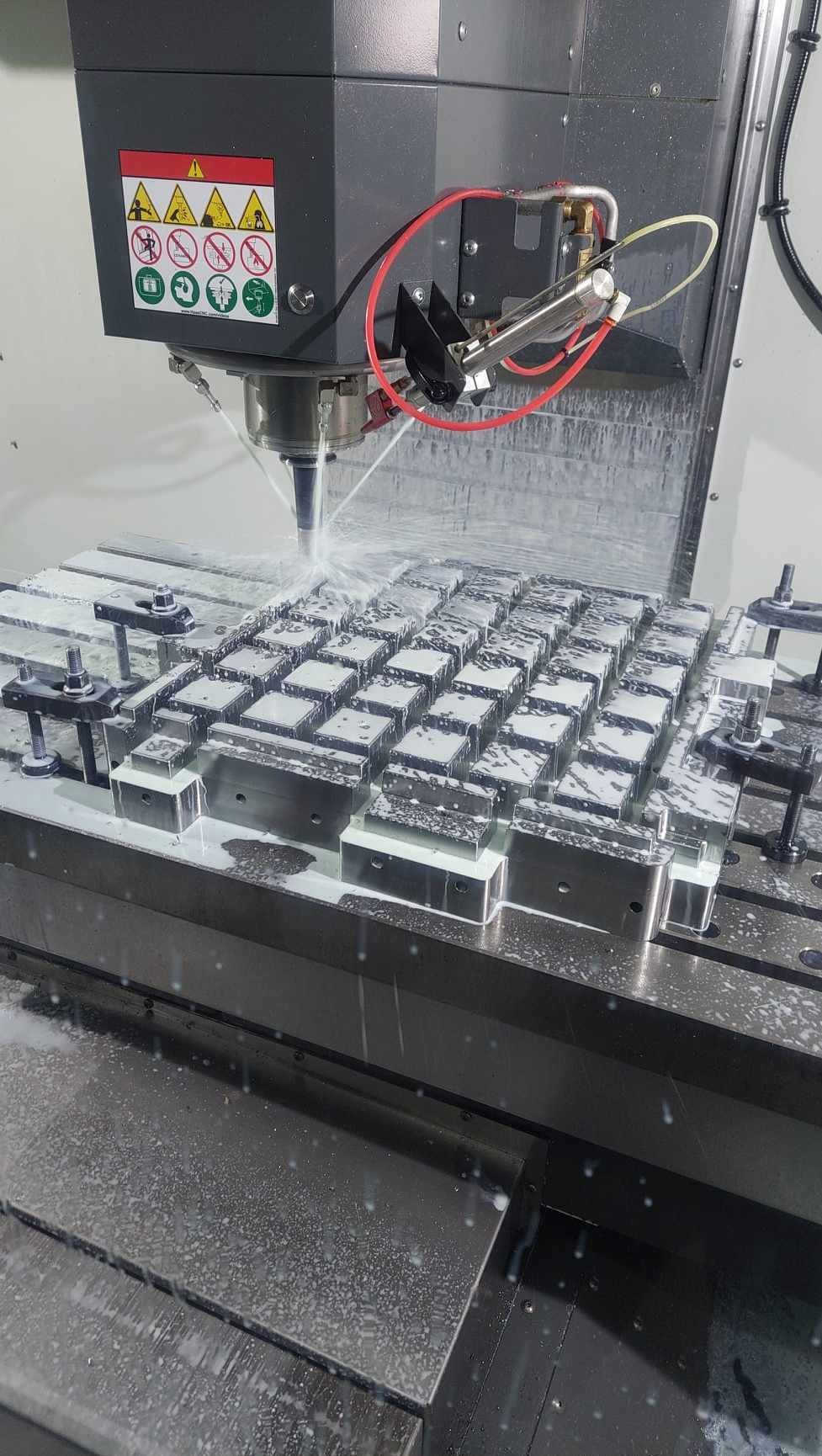 Precyzyjna obróbka metalu na obrabiarce CNC z chłodziwem, detale przymocowane do stołu roboczego, w tle ostrzeżenia BHP.