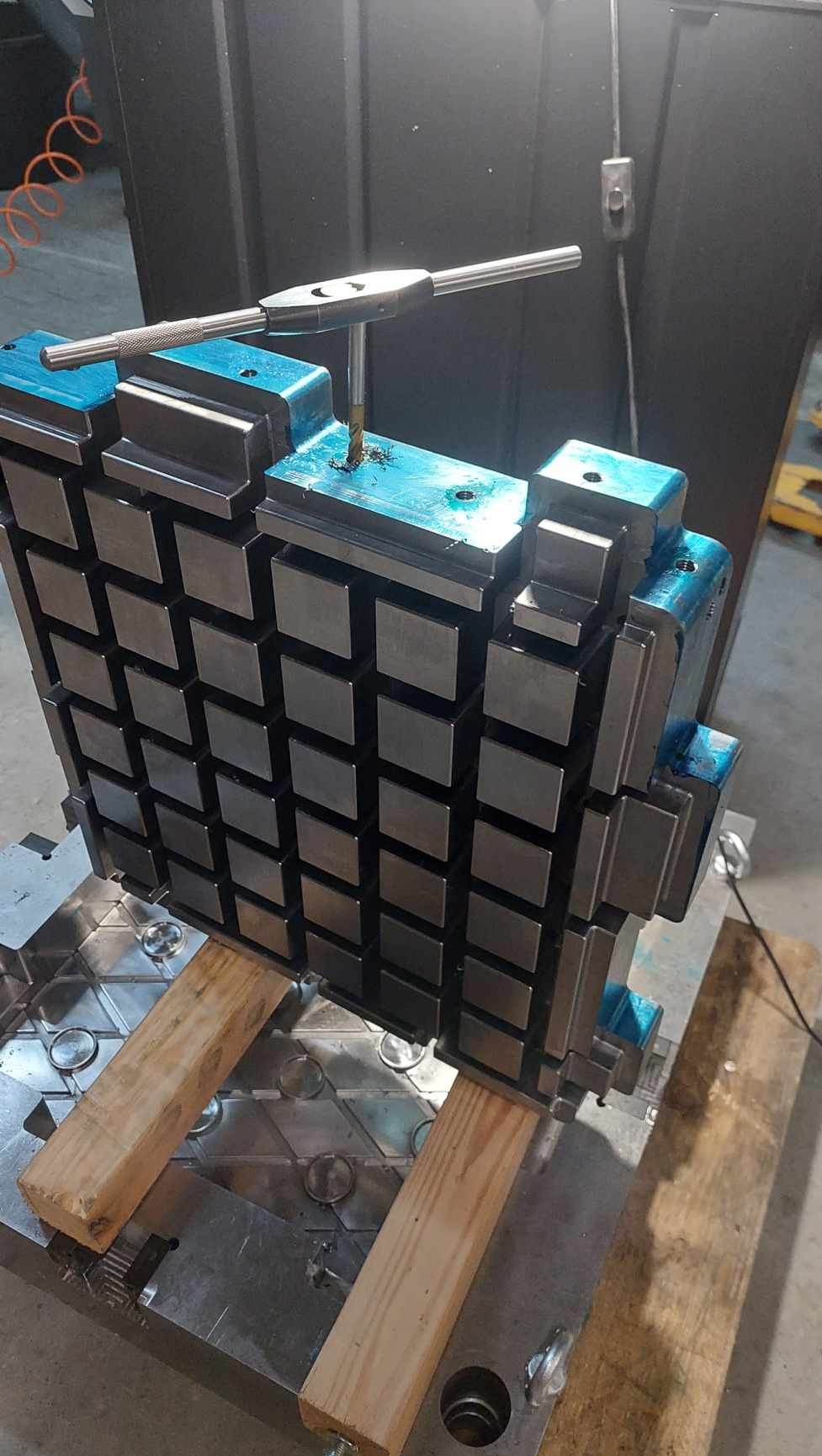 Obróbka metalu: gwintowanie otworów w dużej, modułowej formie z metalu, leżącej na drewnianych legarach. Widoczne wióry i narzędzie do gwintowania.