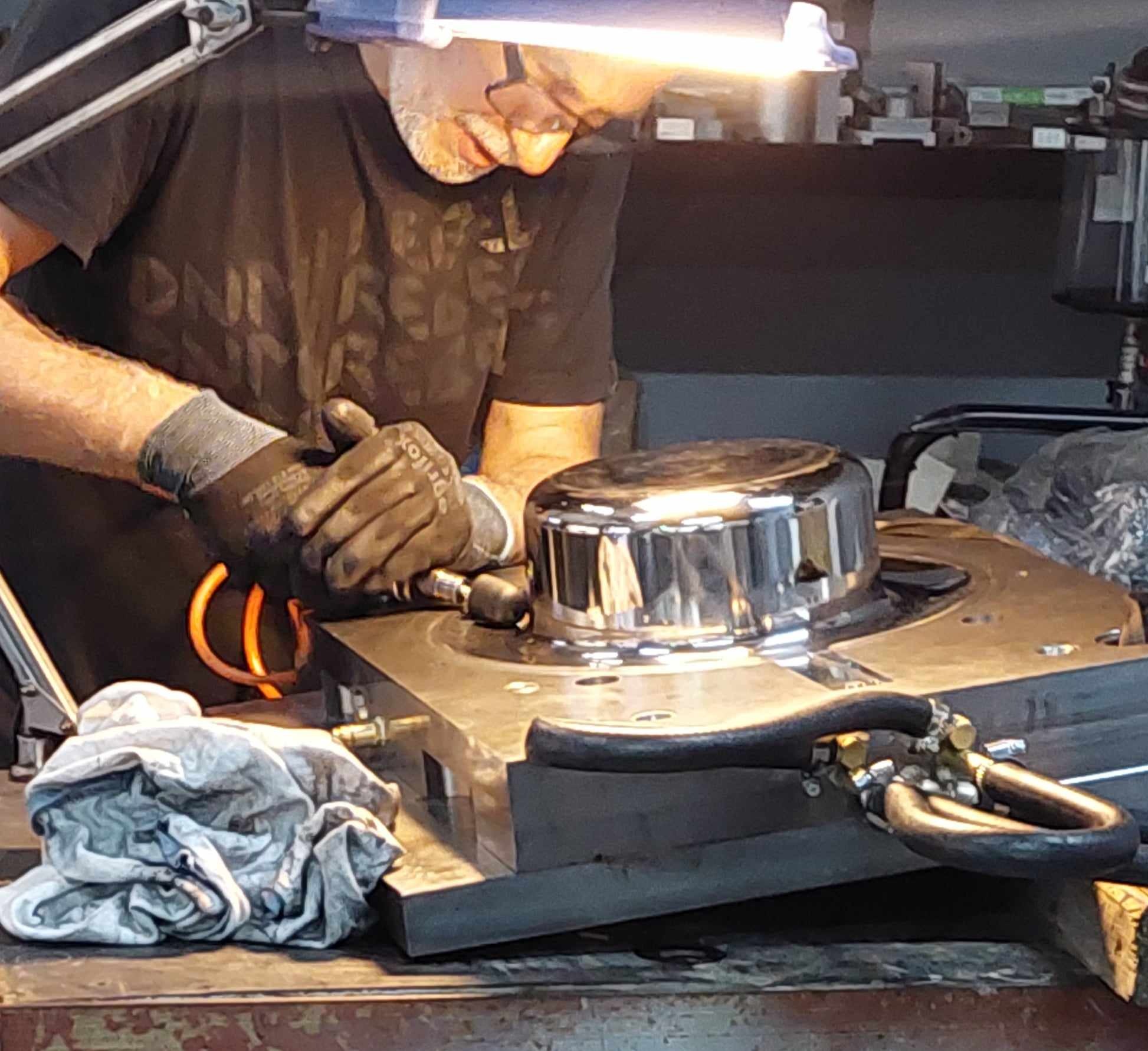 Pracownik w rękawicach poleruje metalowy element na warsztacie. Widoczne narzędzia i lampa oświetlająca. Detale precyzyjnej obróbki metalu w bliskim ujęciu.