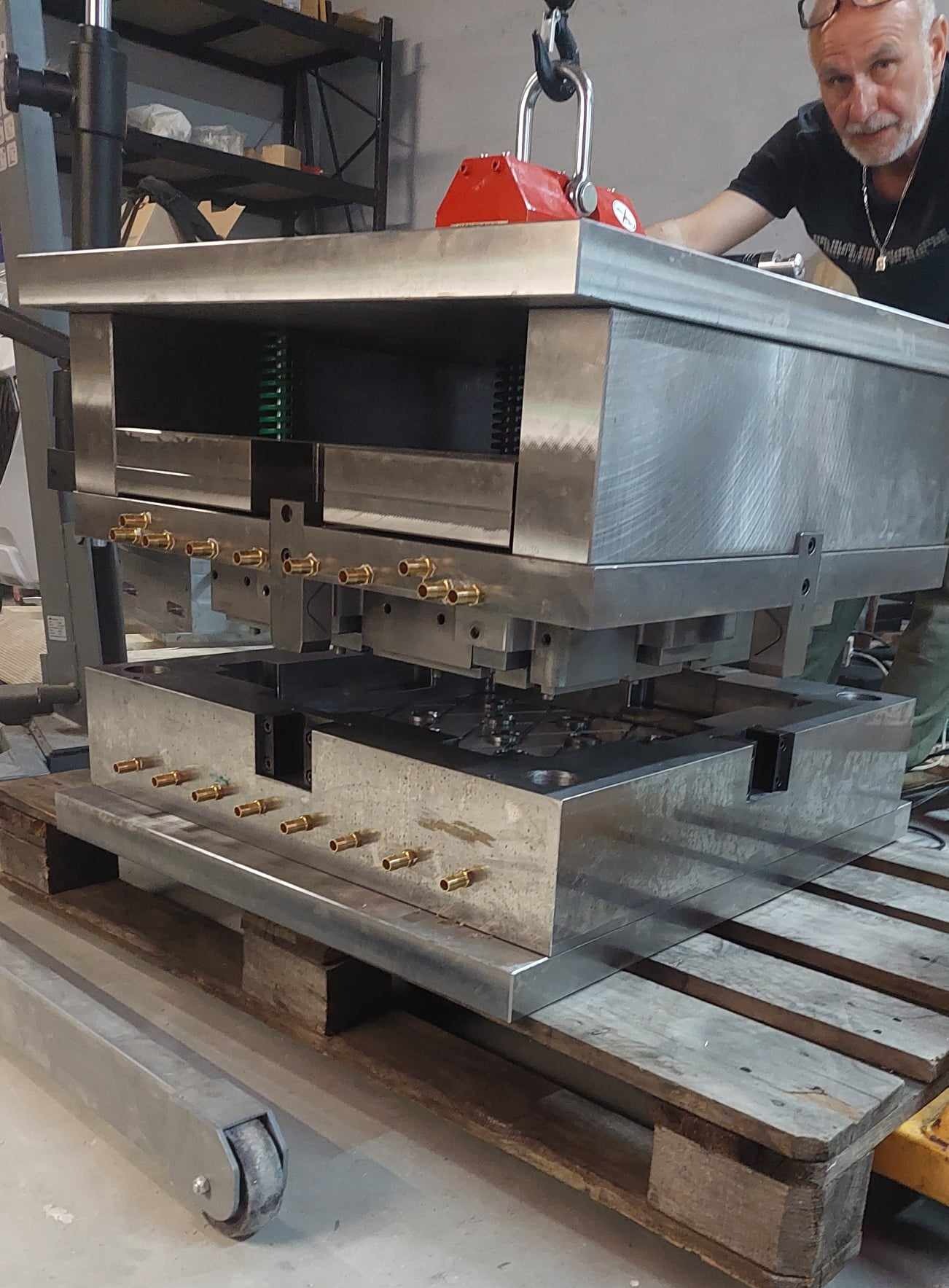 Ciężka, metalowa forma w trakcie montażu z pomocą suwnicy i magnesu, obsługiwana przez mężczyznę w warsztacie. Widoczne elementy hydrauliczne i konstrukcyjne.