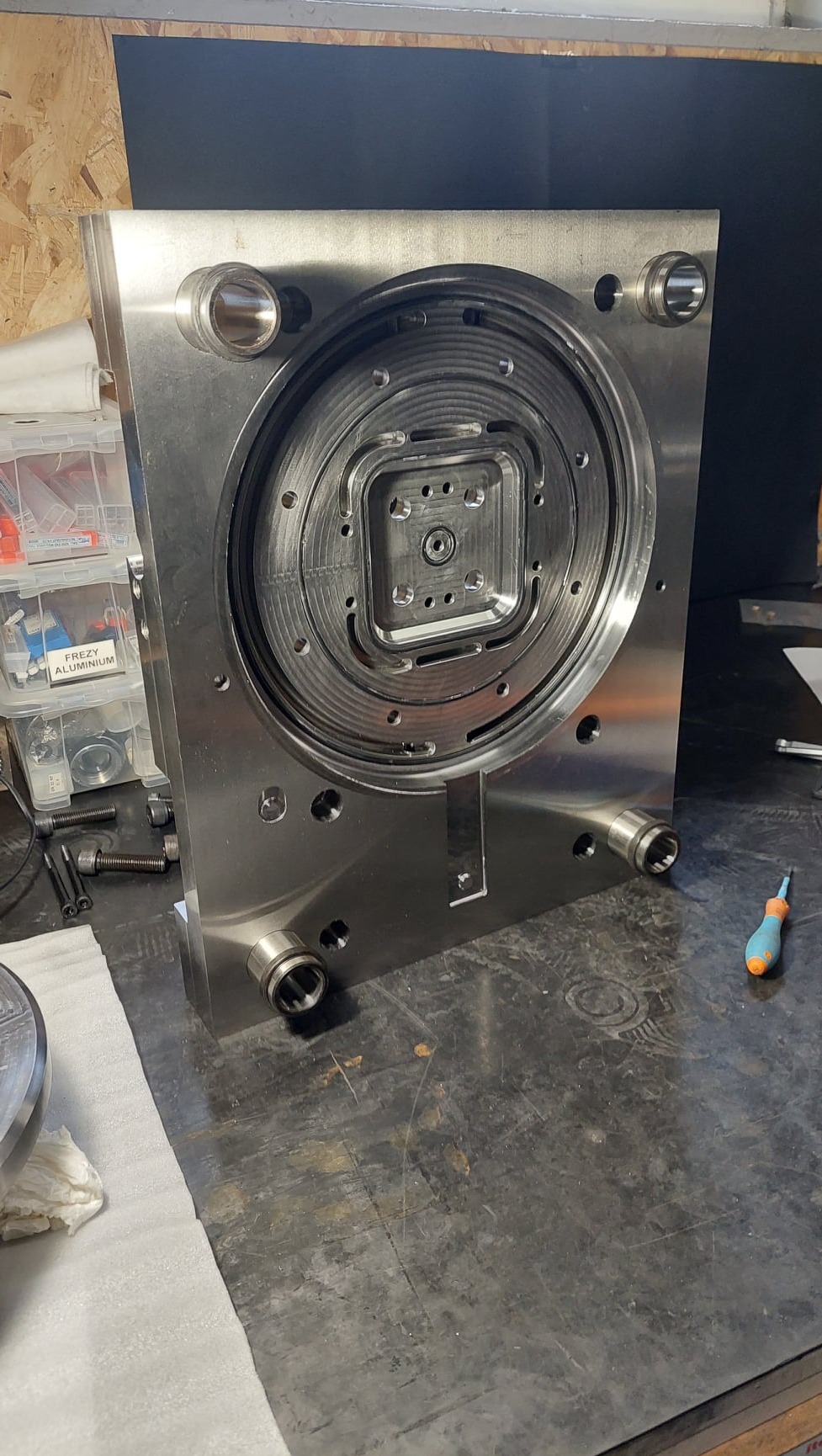 Szczegółowe ujęcie metalowej formy wtryskowej z widocznymi otworami i precyzyjnymi wycięciami, leżącej na warsztatowym stole obok śrubokręta.