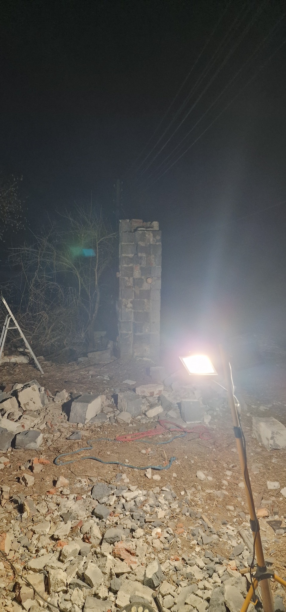 Resztki ceglanego komina w nocnym oświetleniu budowlanym, gruzowisko po rozbiórce, drabina oparta o drzewo w tle.