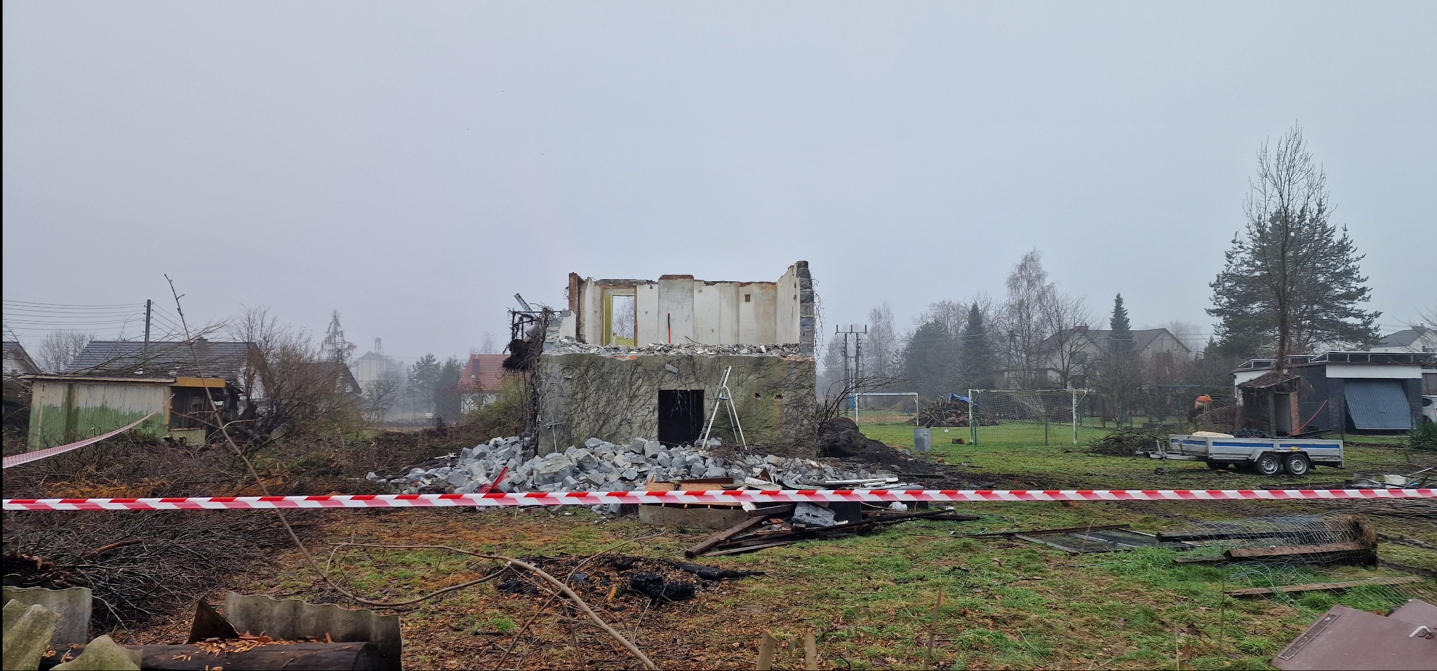Rozbiórka budynku w toku: fragment ściany, gruz, drabina oparta o mur, taśma ostrzegawcza, zieleń w tle. Widok na teren po częściowej rozbiórce w pochmurny dzień.