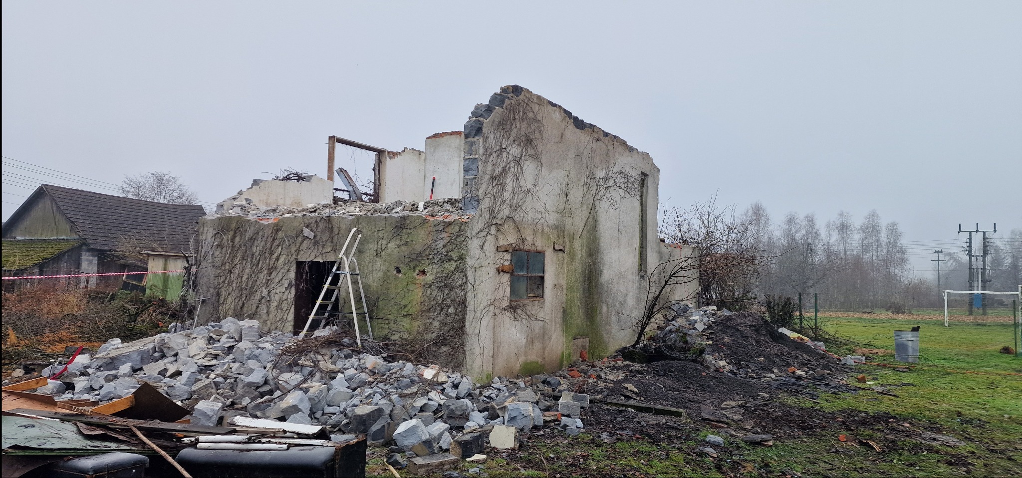 Zrujnowany budynek z resztkami ścian porośniętych bluszczem, gruzowisko i drabina oparta o ścianę. W tle widoczny inny budynek i drzewa w pochmurny dzień.