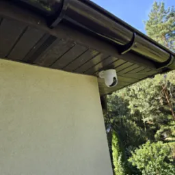 Biała kamera monitoringu zamontowana pod brązowym okapem budynku z beżową elewacją. W tle drzewa i błękitne niebo.