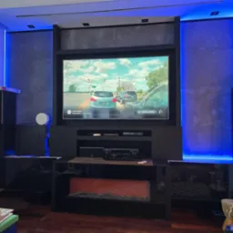 Nowoczesna ściana TV z podświetleniem LED, szarymi panelami i czarnymi szafkami. Na ekranie widoczny monitoring z kamery samochodowej. Elementy inteligentnego domu.