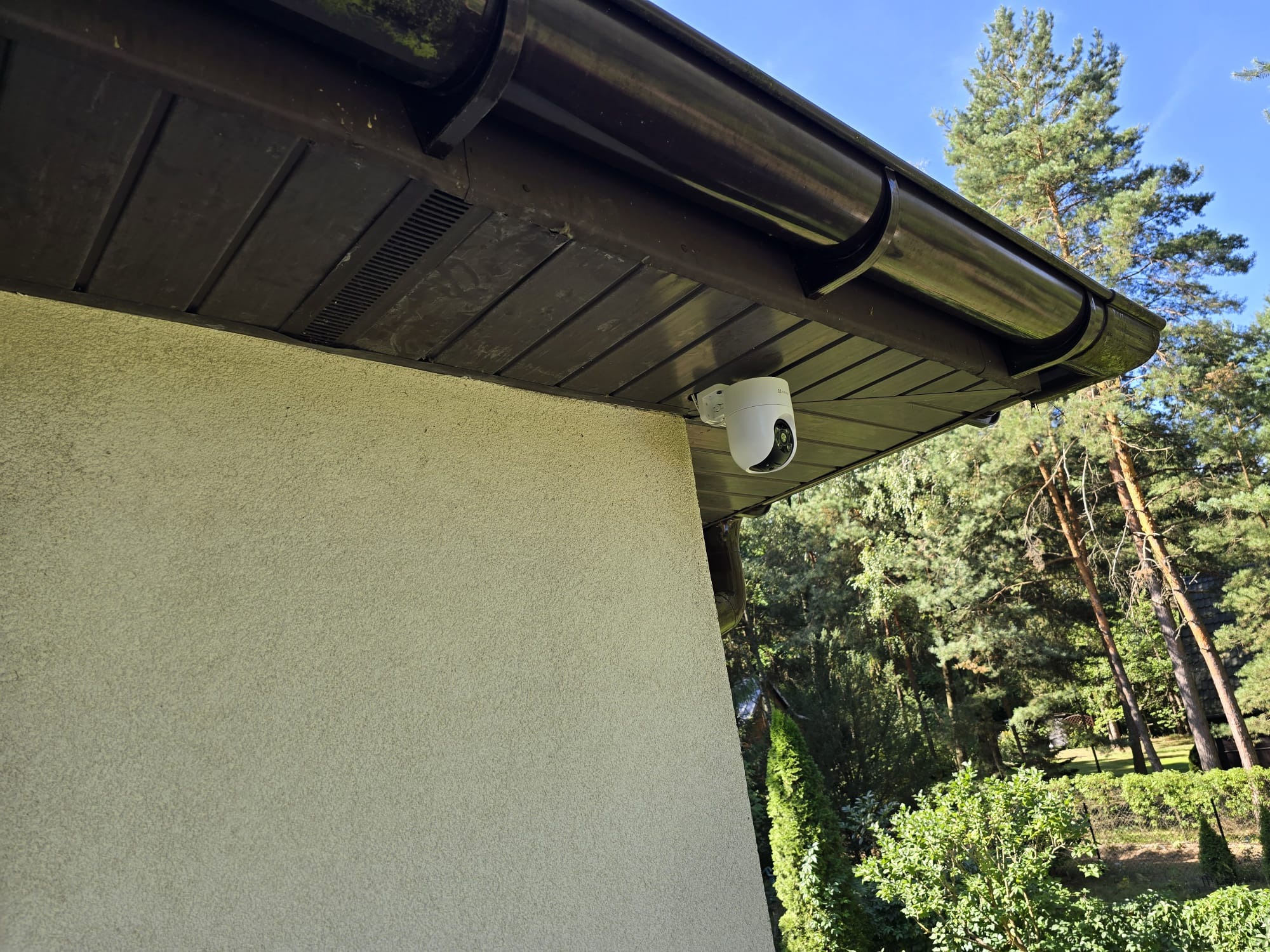 Biała kamera monitoringu zamontowana pod brązowym okapem budynku z beżową elewacją. W tle drzewa i błękitne niebo.