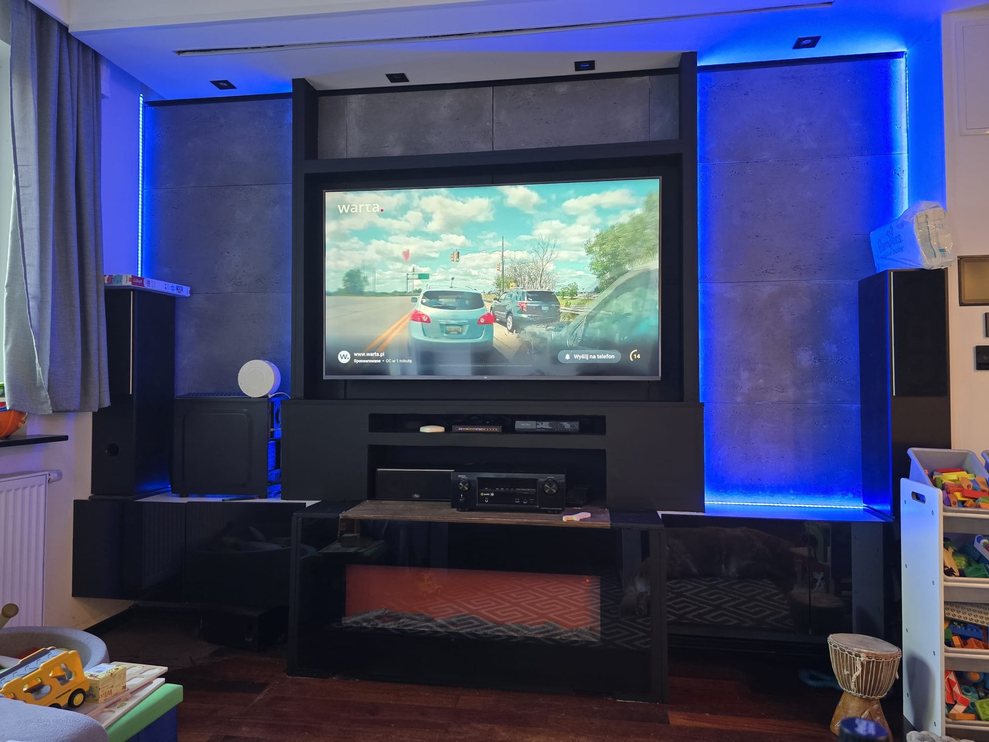 Nowoczesna ściana TV z podświetleniem LED, szarymi panelami i czarnymi szafkami. Na ekranie widoczny monitoring z kamery samochodowej. Elementy inteligentnego domu.