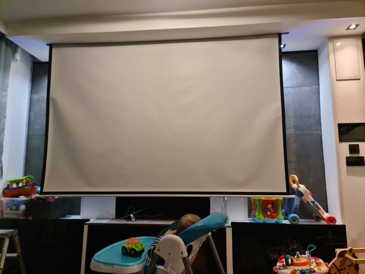 Zasłonięty ekran projekcyjny w pokoju, widoczne zabawki dziecięce. Czarny panel ścienny z szarą strukturą. Biała ściana z włącznikami. Ustawiony w pozycji zwiniętej.