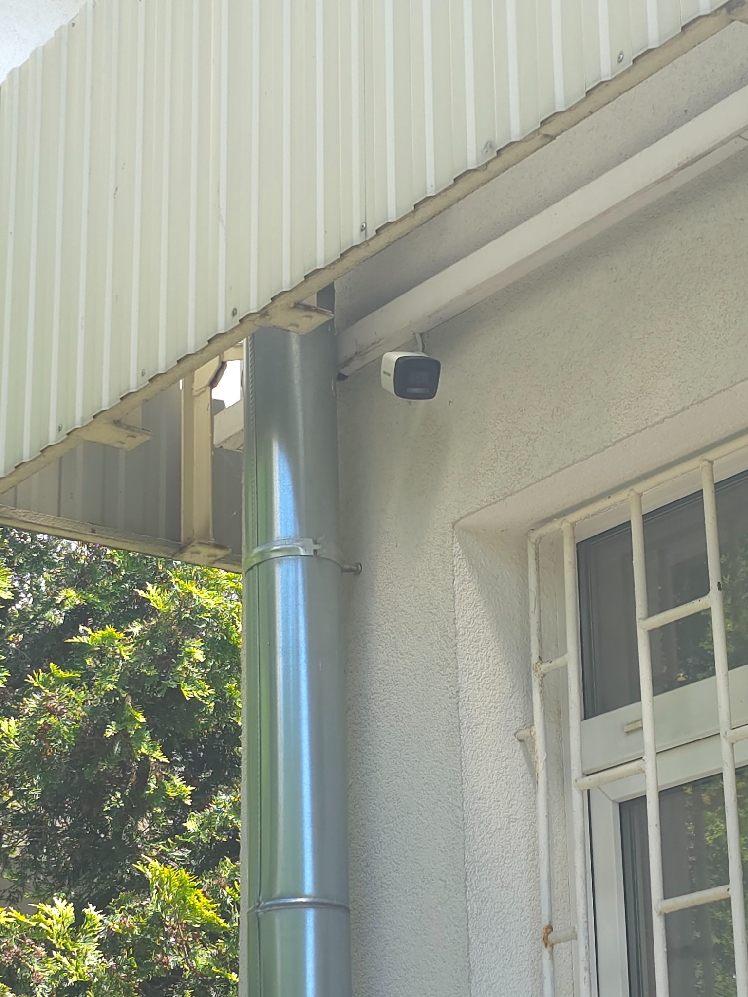 Zewnętrzna kamera monitoringu zamontowana pod okapem budynku, widoczna rynna i okno z kratą. Ujęcie z dołu, w tle zieleń drzew.