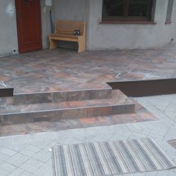 REM-DOM - Wykończony taras z brązowo-szarych płytek imitujących kamień, z trzema schodami i dwoma dywanikami. Widoczny fragment elewacji budynku i zieleń w tle.