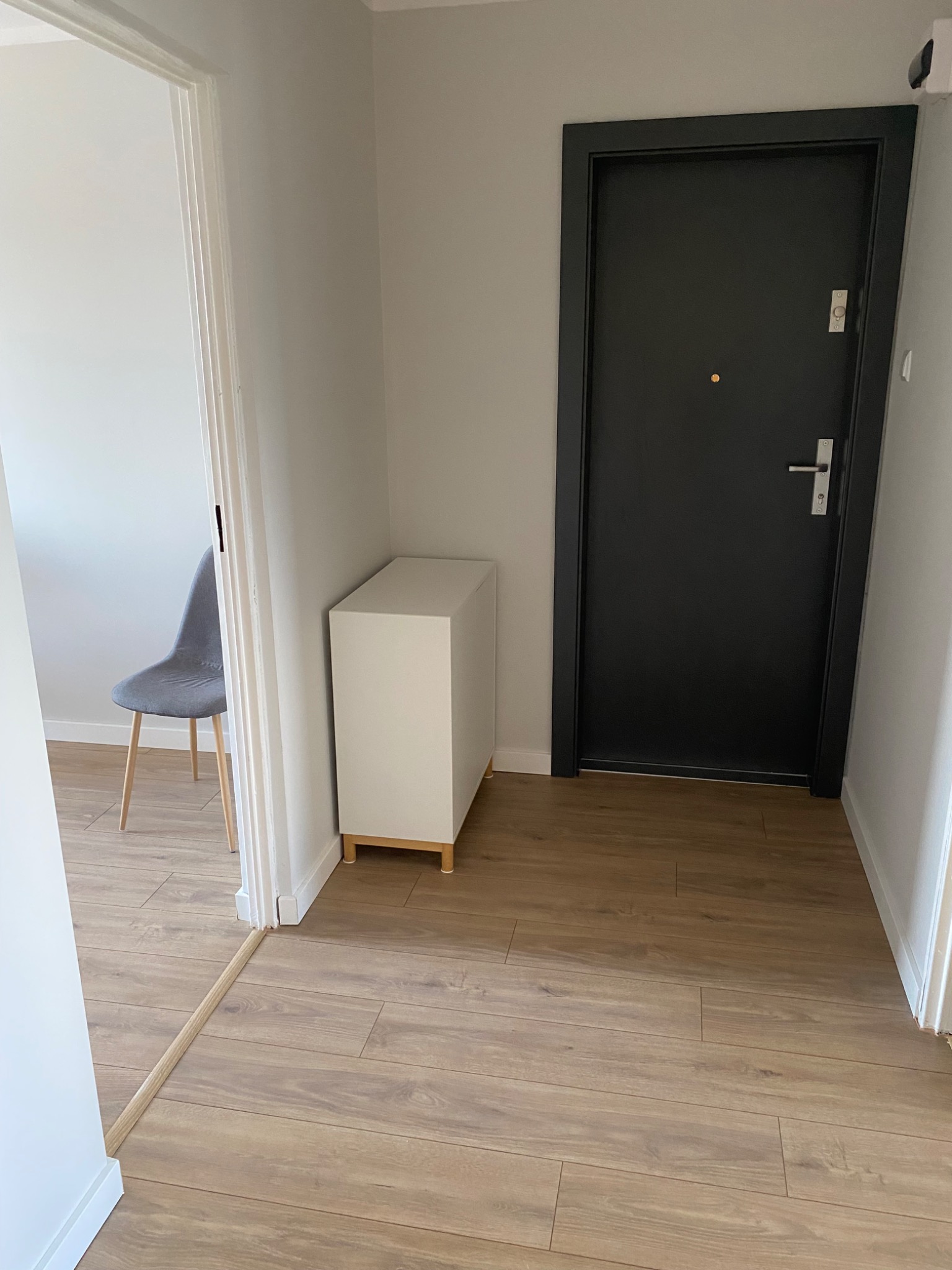 Nowoczesny przedpokój: drewnopodobne panele, biała szafka, czarne drzwi i szare krzesło widoczne w głębi. Minimalistyczny design, jasne ściany.