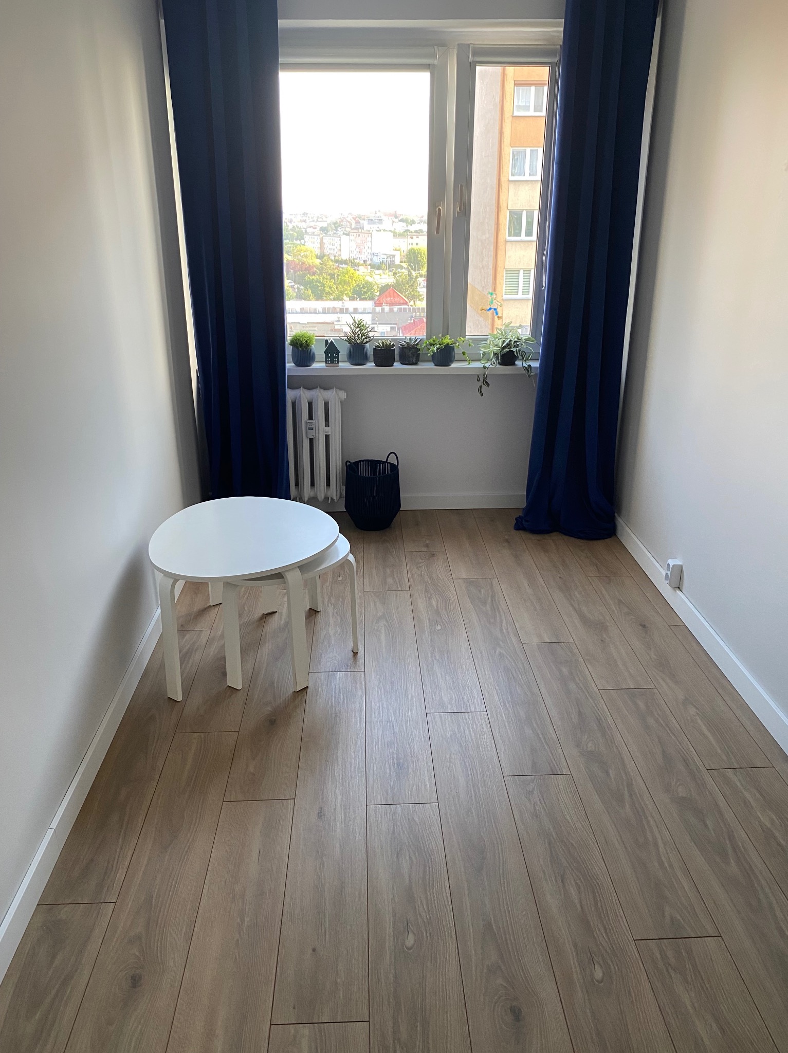 Jasne panele podłogowe w wyremontowanym pokoju z białymi ścianami i niebieskimi zasłonami. Na parapecie rośliny doniczkowe. Widok na miasto.