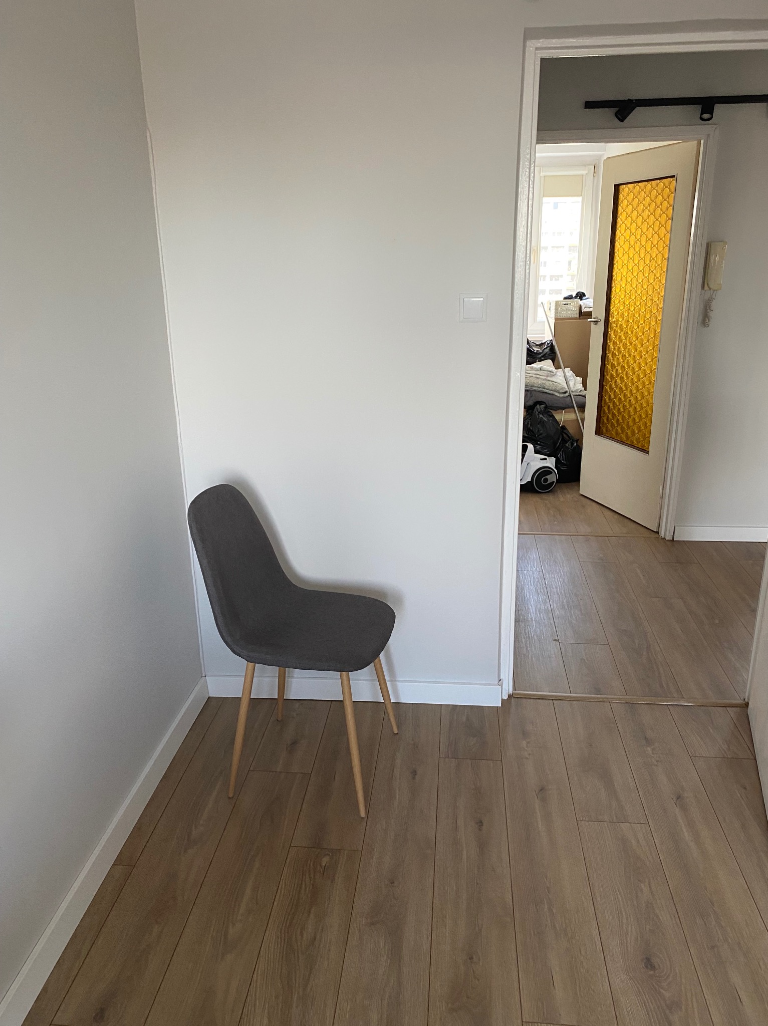 Jasne wnętrze po remoncie: ściany pomalowane na biało, ułożone panele podłogowe, widoczny fragment szarego krzesła i drzwi z żółtą szybą w tle.
