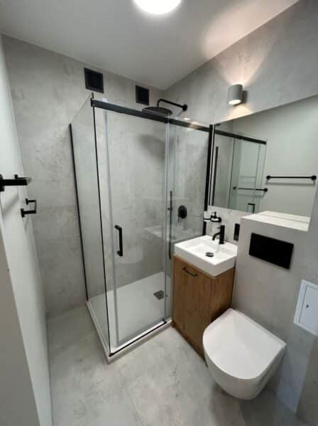 Nowoczesna łazienka z kabiną prysznicową typu walk-in, umywalką na drewnianej szafce i podwieszaną toaletą. Szare płytki na ścianach i podłodze, czarne akcenty.