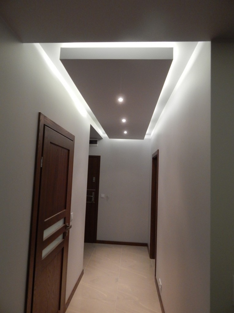 Nowoczesny korytarz z podwieszanym sufitem, oświetleniem LED i drewnianymi drzwiami. Minimalistyczny design, jasne ściany, płytki podłogowe w odcieniach beżu.