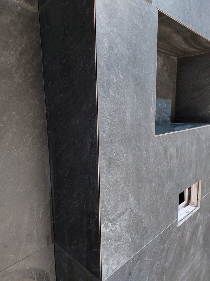 Wykończona ściana z szarych, matowych płytek imitujących kamień, z wnęką i otworem na instalację. Minimalistyczny design łazienki.