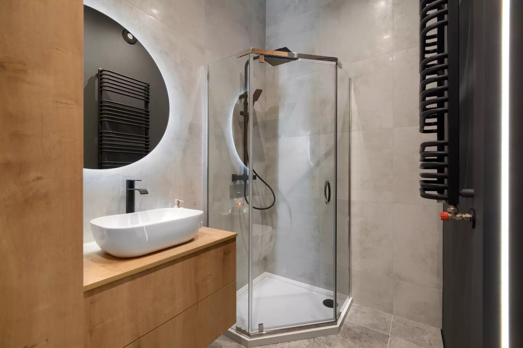 Nowoczesna łazienka z kabiną prysznicową, umywalką na drewnianej szafce i okrągłym lustrem z podświetleniem LED. Minimalistyczny design, szare płytki i czarne akcenty.
