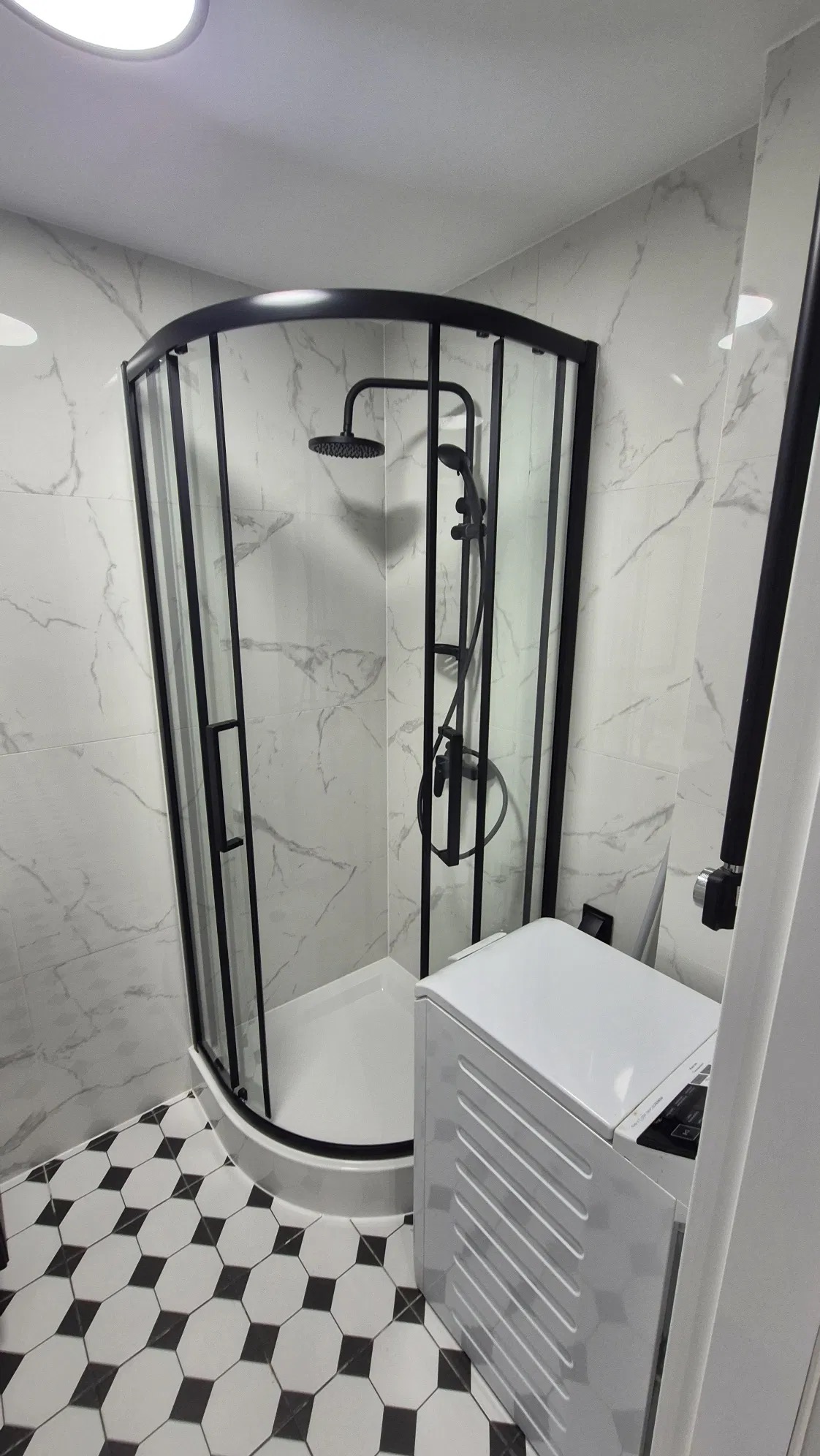 Nowoczesna kabina prysznicowa z czarnymi ramami w łazience wykończonej marmurowymi płytkami i wzorzystą podłogą, obok pralka.