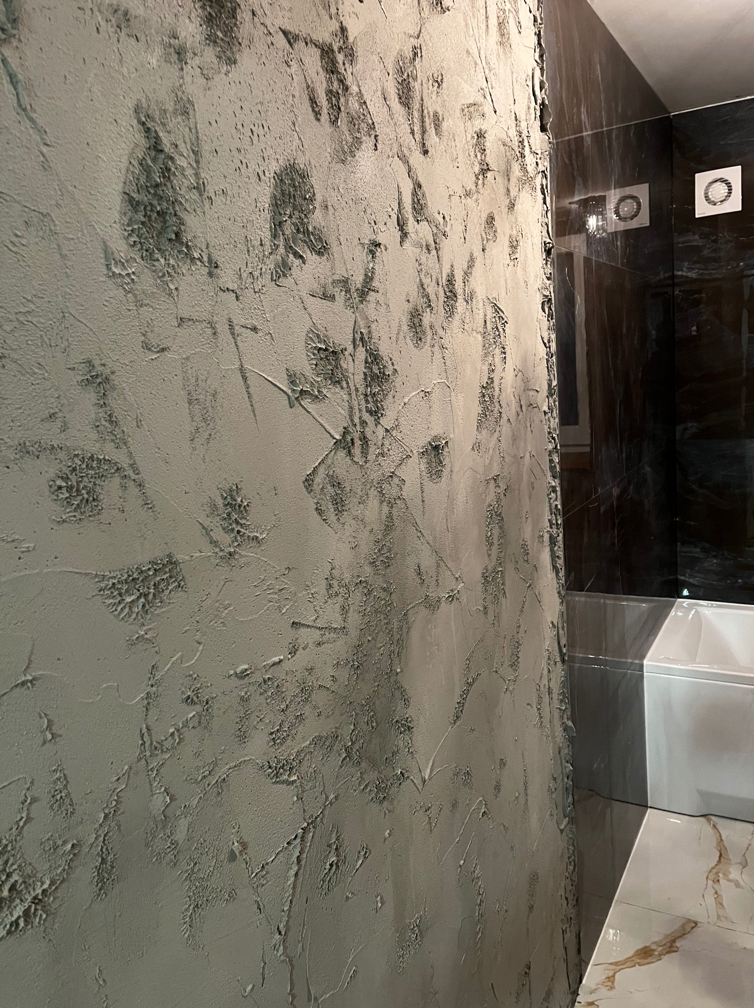Ściana w łazience z nałożoną szpachlą dekoracyjną, widoczne nierówności i faktura. Obok fragment czarnej ściany z marmurowym wzorem i biała wanna.