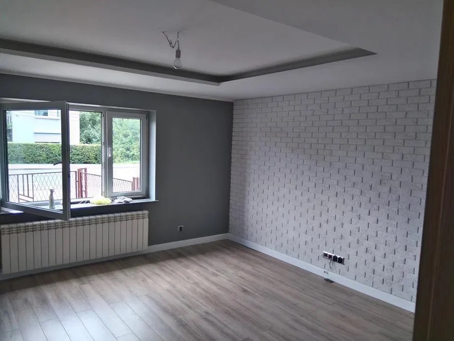 Pokój po remoncie: szara ściana, biała ściana z cegły, drewniana podłoga, otwarte okno z widokiem na zieleń, grzejnik i sufit z podwieszanym oświetleniem.