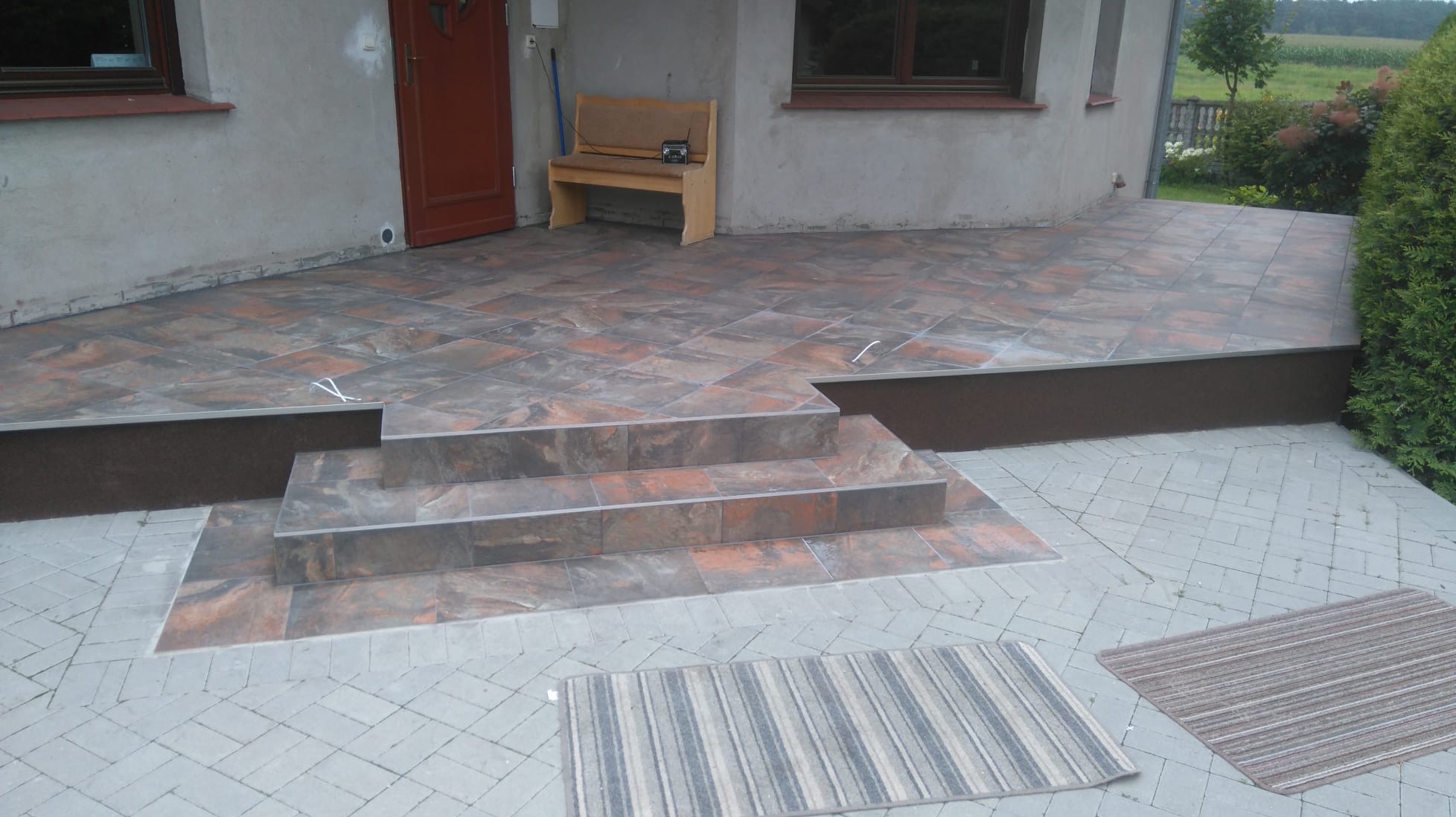 Wykończony taras z brązowo-szarych płytek imitujących kamień, z trzema schodami i dwoma dywanikami. Widoczny fragment elewacji budynku i zieleń w tle.