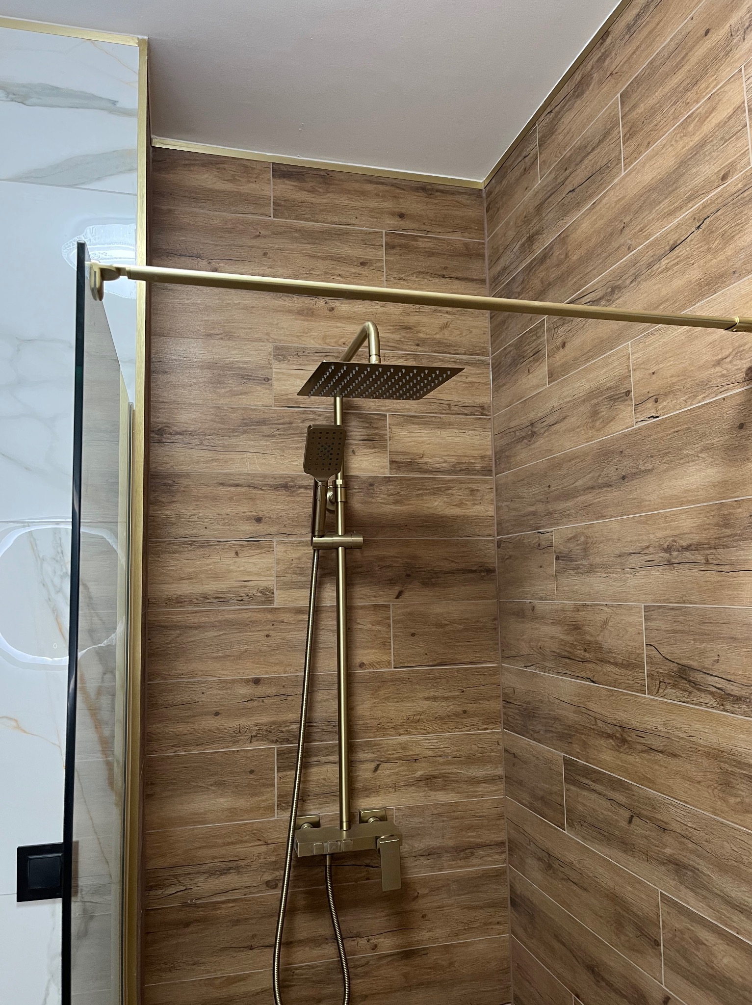 Nowoczesna kabina prysznicowa z drewnopodobnymi płytkami i złotą armaturą, widok z bliska na deszczownicę i baterię.