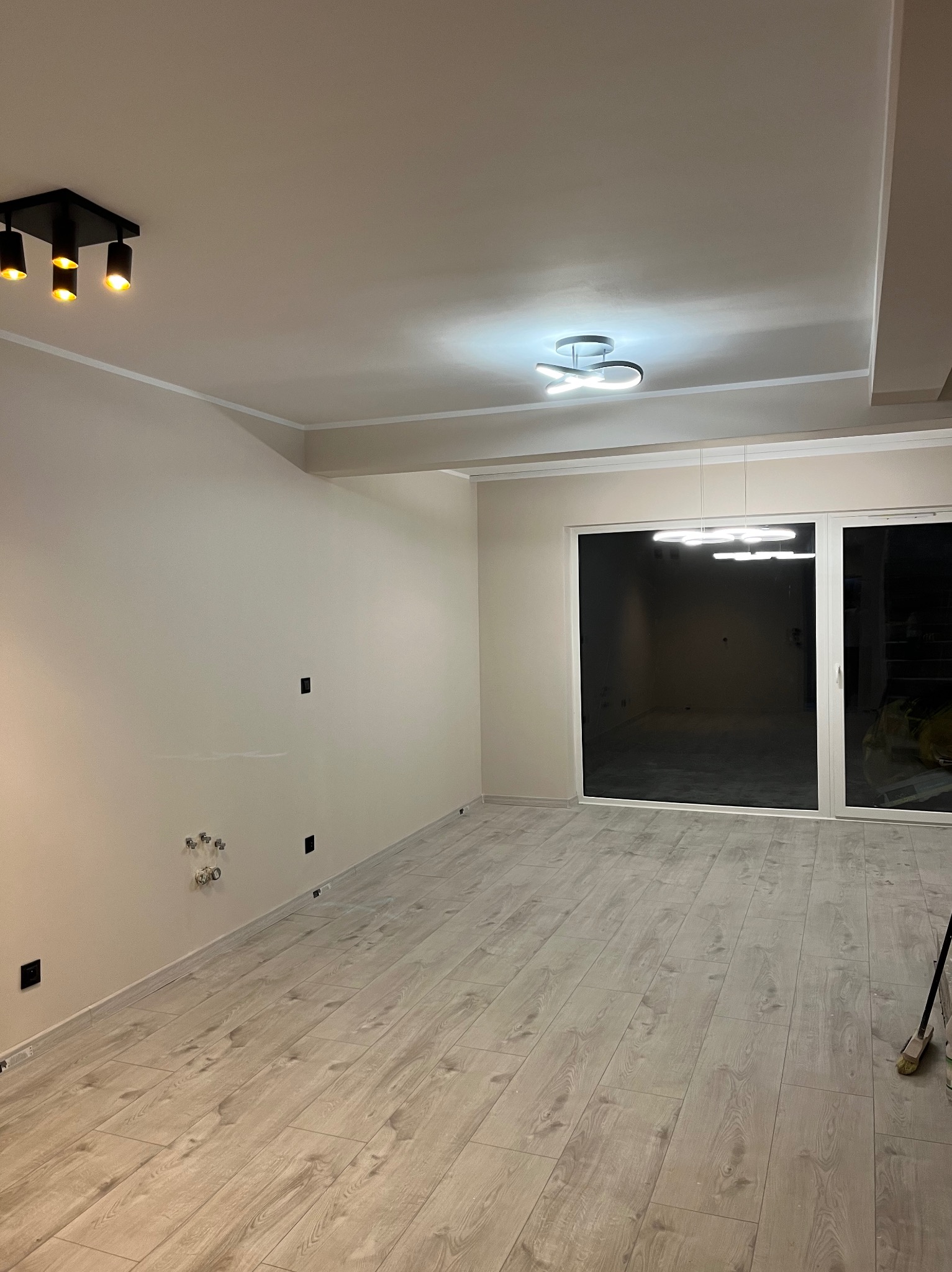 Jasne panele podłogowe w nowym, przestronnym pokoju z beżowymi ścianami, nowoczesnym oświetleniem sufitowym i dużym oknem balkonowym. Widoczne gniazdka elektryczne.