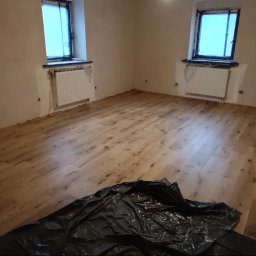 MAYSTER - Jasna, drewniana podłoga po renowacji w pokoju z dwoma oknami. Grzejniki zabezpieczone folią, widoczna czarna plandeka ochronna na podłodze.