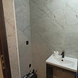 MAYSTER - Wykończona łazienka z marmurowymi płytkami, umywalką z czarną armaturą i narzędziem glazurniczym w rogu. Widoczne gniazdka elektryczne.