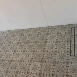 Ukończona podłoga w łazience z szarymi, wzorzystymi płytkami, widoczny odpływ liniowy i fragment ściany z surowym wykończeniem. Biały sufit z fugami.