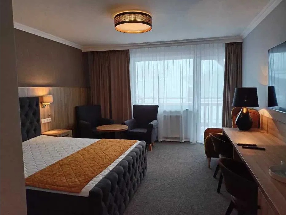 Elegancko wykończony pokój hotelowy z dużym łóżkiem, stolikiem, krzesłami i balkonem, utrzymany w stonowanej kolorystyce brązów i szarości, oświetlony ciepłym światłem.