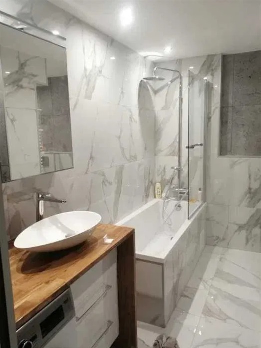 Nowoczesna łazienka z wanną i umywalką nablatową. Białe płytki imitujące marmur na ścianach i podłodze. Drewniany blat pod umywalką.