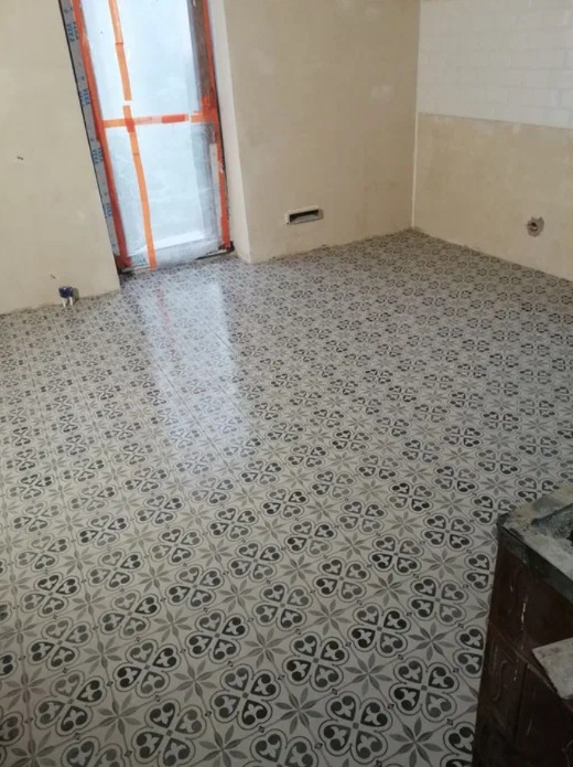 Ukończona podłoga w kuchni wyłożona wzorzystymi, szaro-białymi płytkami. Widoczna ściana z fragmentem białej glazury i okno z zabezpieczeniami remontowymi.