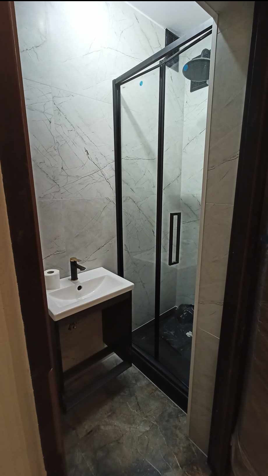 Nowoczesna łazienka z kabiną prysznicową w czarnej ramie i umywalką na szafce. Marmurowe płytki ścienne, szara posadzka. Wnętrze po remoncie, widok zza drzwi.