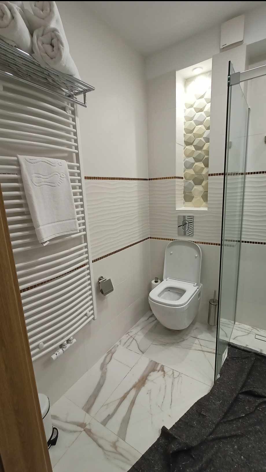 Nowoczesna łazienka z białą armaturą, grzejnikiem drabinkowym z ręcznikami, płytkami z efektem 3D i mozaiką. Marmurowe płytki podłogowe.