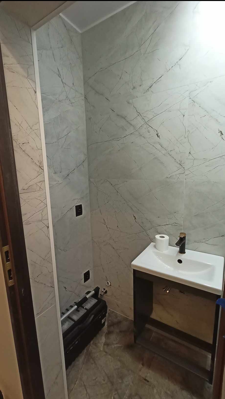 Wykończona łazienka z marmurowymi płytkami, umywalką z czarną armaturą i narzędziem glazurniczym w rogu. Widoczne gniazdka elektryczne.