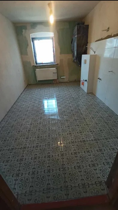 Wykończona podłoga z płytek w łazience w trakcie remontu. Widoczne okno, grzejnik i fragment instalacji. Ściany częściowo przygotowane pod malowanie.