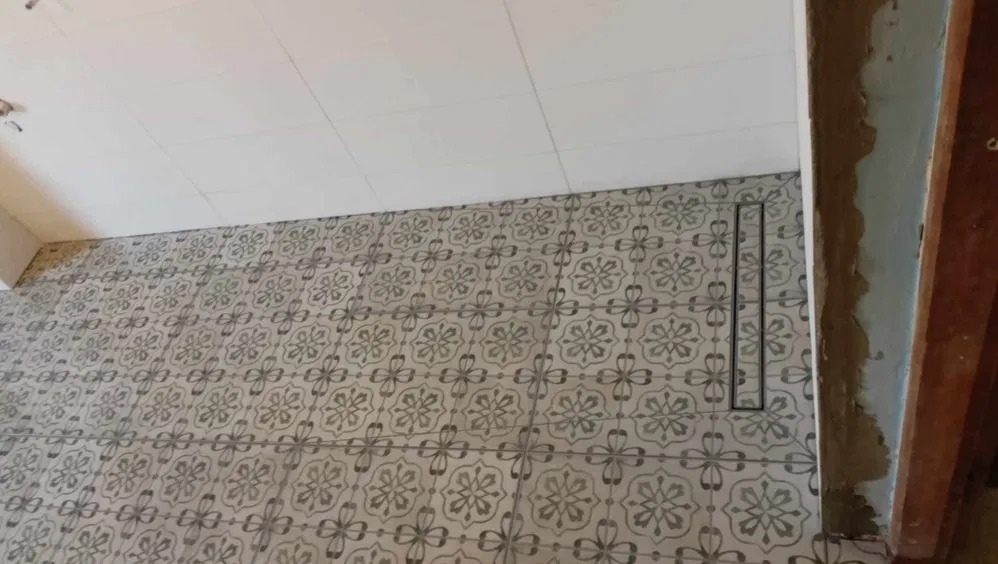 Ukończona podłoga w łazience z szarymi, wzorzystymi płytkami, widoczny odpływ liniowy i fragment ściany z surowym wykończeniem. Biały sufit z fugami.