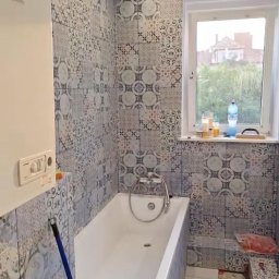 Adnabud - Wykończona łazienka z wanną, zdobiona mozaiką w odcieniach szarości i błękitu. Widoczny bojler, okno z widokiem na zieleń i fragment podłogi z białych płytek.