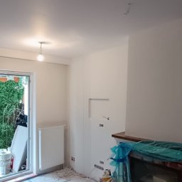 Adnabud - Wnętrze w trakcie malowania: świeżo pomalowane ściany w odcieniach bieli, zabezpieczony kominek folią, okno zasłonięte, widoczne ślady prac malarskich na podłodze.