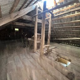 Adnabud - Stare, drewniane poddasze z widoczną konstrukcją dachu i podłogi. Fragmenty desek podłogowych są uszkodzone z dziurami. Widoczne drewniane podpory i niewielkie okno.