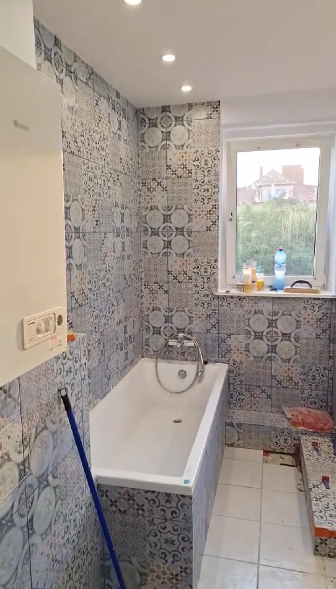 Wykończona łazienka z wanną, zdobiona mozaiką w odcieniach szarości i błękitu. Widoczny bojler, okno z widokiem na zieleń i fragment podłogi z białych płytek.