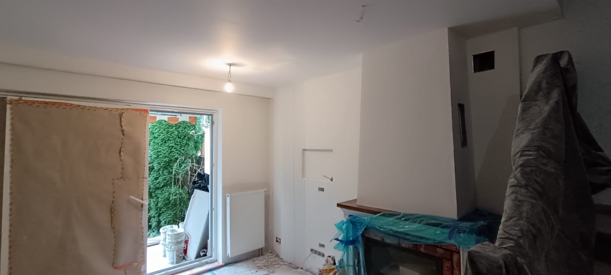 Wnętrze w trakcie malowania: świeżo pomalowane ściany w odcieniach bieli, zabezpieczony kominek folią, okno zasłonięte, widoczne ślady prac malarskich na podłodze.