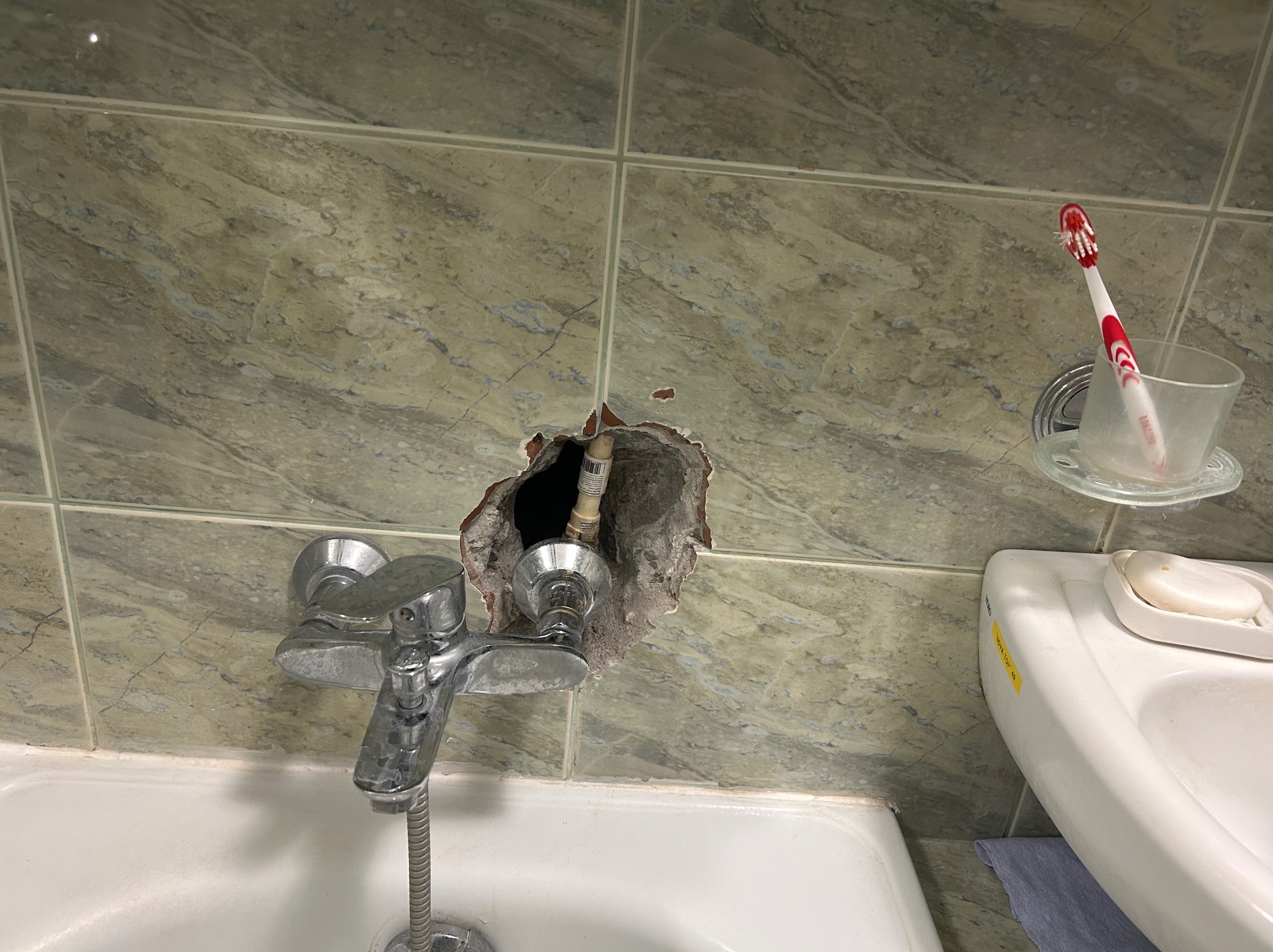 Uszkodzona ściana z płytek w łazience, z widocznym otworem wokół rury wodociągowej, bateria umywalkowa i akcesoria łazienkowe. Konieczna naprawa.