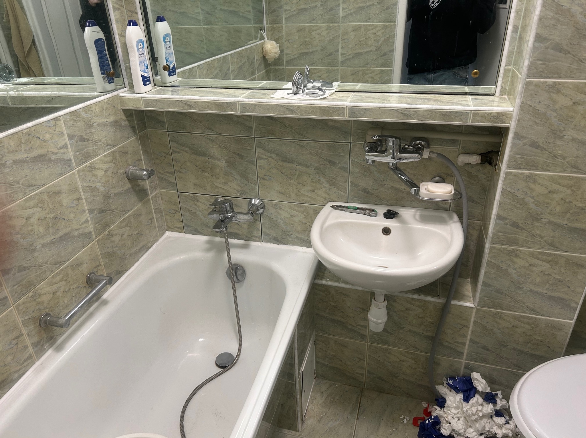 Umywalka i wanna w łazience wyłożonej płytkami w odcieniach zieleni. Na umywalce leży nóż do tapet. Widoczne akcesoria łazienkowe i fragment toalety.