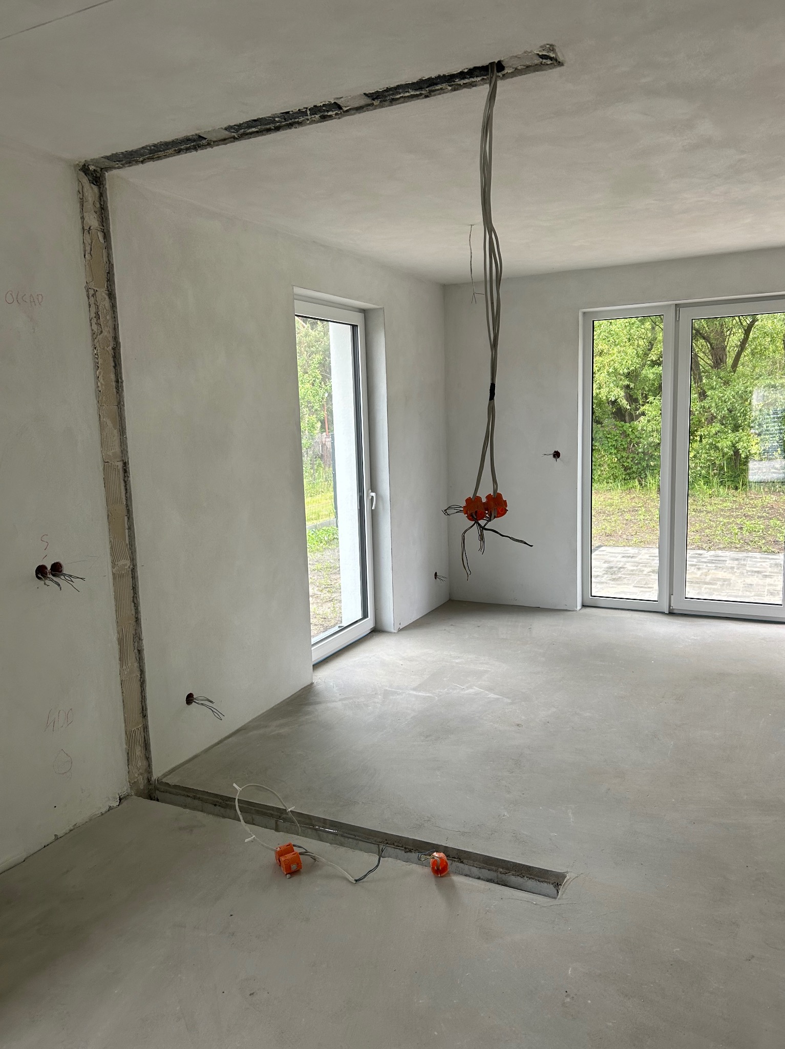 Wnętrze w trakcie remontu: szpachlowane ściany, wnęki pod instalacje elektryczne, kable zwisające z sufitu, wylewka podłogowa. Widoczne okna z widokiem na zieleń.